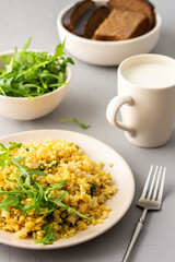 Delicious bulgur porridge with fried onions and herbs in a plate, a glass of milk, arugula in a bowl and black bread