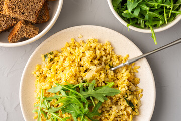 Delicious bulgur porridge with fried onions and herbs in a plate, arugula in a bowl and black bread
