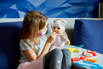 A little girl in medical mask sits on the sofa, plays a doctor, treats to the doll