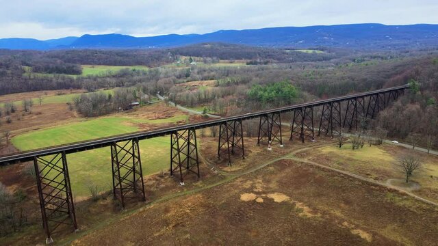 Aerial drone video footage of a train bridge viaduct running over a valley in the Appalachain Mountains during early spring on a cloud day, surrounded by mountains and farmland. 