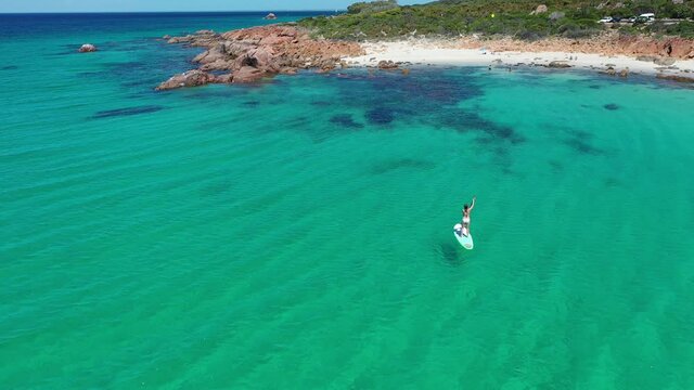 Aerial view of female standing on a paddle board in turquoise sea water by white sand beach of Point Picquet, Cape Naturaliste, Australia