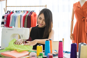 Smiling Happy Asian woman using a sewing machine to sew on clothe. Lady is working owner sketch dressmaker for customers in tailor shop. Concept designer maker clothing.