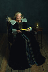 Senior woman dressed in Renaissance attire with a book sitting in a chair by a small table topped by a candlestick near a dark wall.
