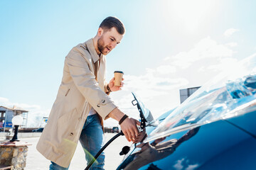 Stylish man with coffe cup in hand inserts plug into the electric car charging socket