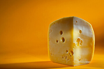 Yellow piece of cheese on an orange background. Triangular cheese close up. Copy space and free space for text near the cheese. Contrasting light as an artistic effect.