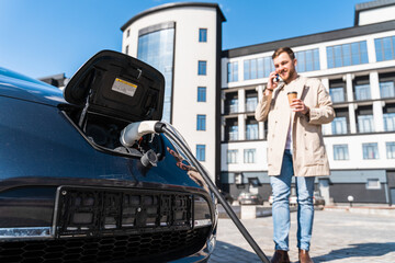 Obraz premium Stylish man talks on the phone and drinks coffee while his car is charging at the station