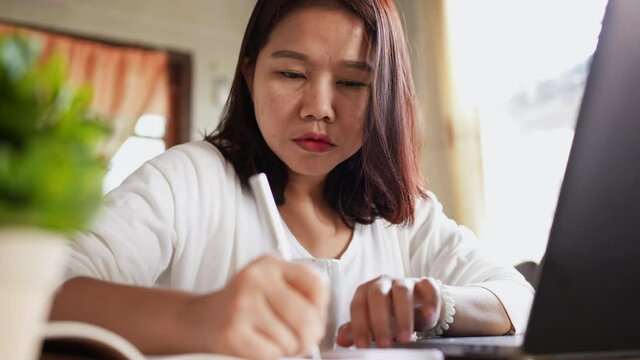 close up on young asian woman feeling headache while writing list of expense after calculated about money by calculator at desk with financial bill and document ,millennial lifestyle with unhealthy fi