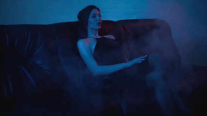 brunette woman in dress sitting on black couch on blue with smoke