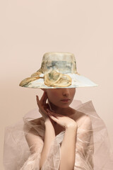 Mystery lady with half hidden face is wearing beige brim hat made of organza with floral design and decorated with cloth bow. Romantic woman in mesh wrap is posing on the beige background. 