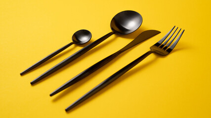  Black utensils set on yellow background. Black kitchen tableware. Stylish metal spoon, teaspoon, fork and knife.