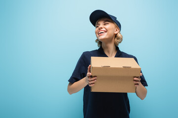 cheerful courier laughing while holding carton package isolated on blue