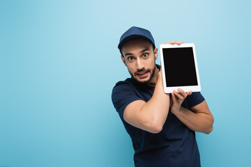 thrilled arabian courier looking at camera while holding digital tablet with blank screen isolated on blue