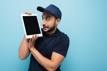 shocked arabian courier looking at digital tablet with blank screen isolated on blue