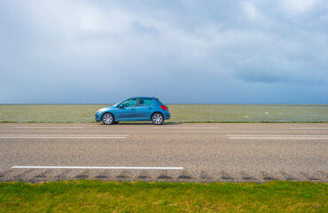 Obraz na płótnie Canvas Car on a dike defying a stormy lake below a blue sky and white gray clouds in spring, Almere, Flevoland, The Netherlands, April 5, 2021 