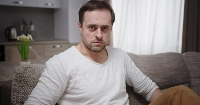 Piercing serious menacing look at camera young depressed man sitting on sofa frustrated showing displeasure