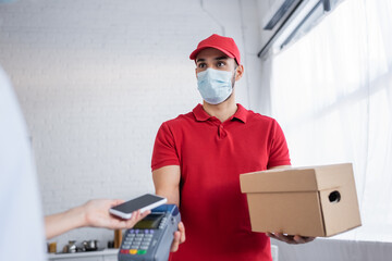 muslim courier in medical mask holding payment terminal near woman with smartphone on blurred foreground