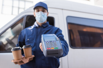 Fototapeta na wymiar selective focus of coffee to go and payment terminal in hands of arabian courier in medical mask on blurred background