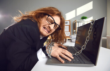 Corporate slave. Side view of a tired crazy office worker who works overtime chained to a laptop and cries. Concept of workload, extra hours and modern corporate slavery.