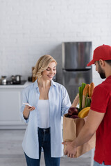 smiling woman holding smartphone near arabian courier with fresh food in paper bag