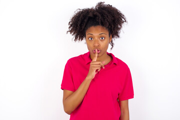 young beautiful African American woman wearing pink t-shirt against white wall makes hush gesture, asks be quiet. Don't tell my secret or not speak too loud, please!