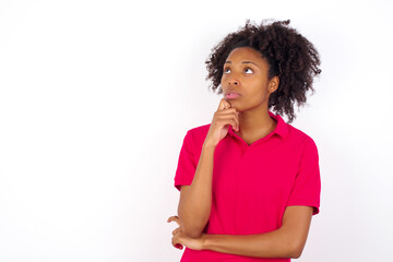 Fototapeta na wymiar Portrait of thoughtful young beautiful African American woman wearing pink t-shirt keeps hand under chin, looks away trying to remember something or listens something with interest. Youth concept.