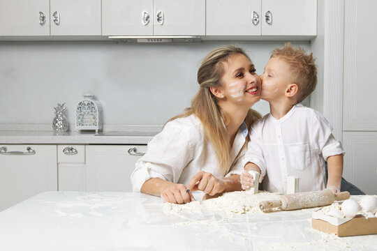 Mom and son were cooking from dough in a white kitchen and smeared with flour, the boy kisses his mother