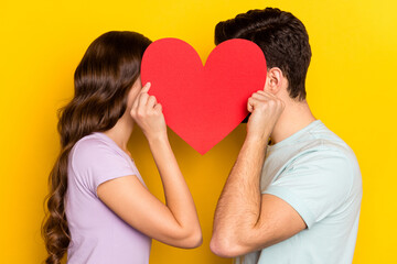 Profile side view portrait of couple holding big heart hiding face romance kissing isolated over bright yellow color background