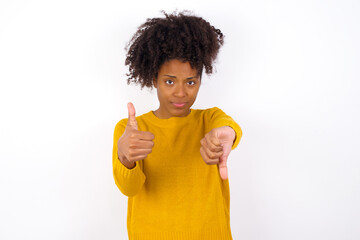 young beautiful African American woman wearing yellow sweater against white wall feeling unsure making good bad sign. Displeased and unimpressed.