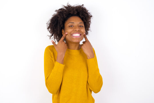 Strong healthy straight white teeth. Close up portrait of happy young beautiful African American woman wearing yellow sweater against white with beaming smile pointing on perfect clear white teeth.