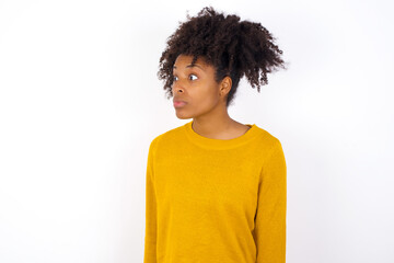Obraz na płótnie Canvas young beautiful African American woman wearing yellow sweater against white wall stares aside with wondered expression has speechless expression. Embarrassed model looks in surprise