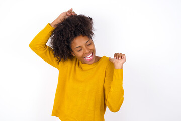 Photo of upbeat young beautiful African American woman wearing yellow sweater against white wall has fun and dances carefree wear being in perfect mood makes movements. Spends free time on disco party