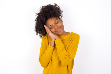 young beautiful African American woman wearing yellow sweater against white wall leans on pressed palms closes eyes and has pleasant smile dreams about something