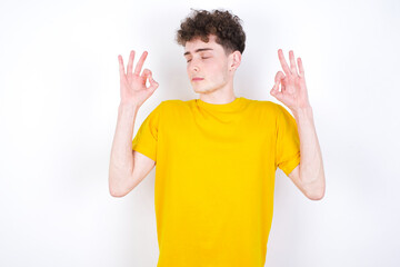 young caucasian handsome man with curly hair wearing yellow T-shirt against white studio background  relax and smiling with eyes closed doing meditation gesture with fingers. Yoga concept.