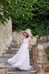 Beautiful woman with purple hair in a white wedding dress walks in the garden. Natural makeup