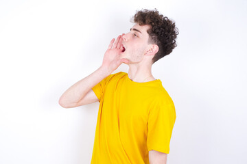 young caucasian handsome man with curly hair wearing yellow T-shirt against white studio background  shouting and screaming loud to side with hand on mouth. Communication concept.