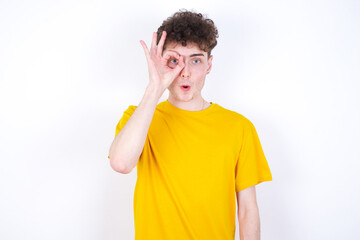 young caucasian handsome man with curly hair wearing yellow T-shirt against white studio background  doing ok gesture shocked with surprised face, eye looking through fingers. Unbelieving expression.
