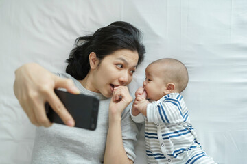 Young mother and newborn baby taking selfie with mobile phone on bed.