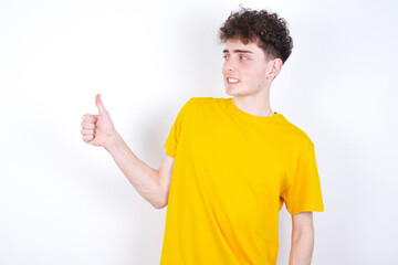 young caucasian handsome man with curly hair wearing yellow T-shirt against white studio background  Looking proud, smiling doing thumbs up gesture to the side. Good job!