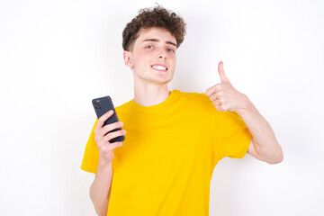 Portrait of young caucasian handsome man with curly hair wearing yellow T-shirt using and texting with smartphone  happy with big smile doing ok sign, thumb up with fingers, excellent sign