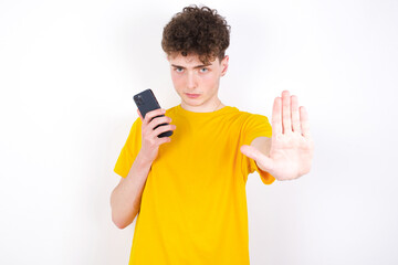 young caucasian handsome man with curly hair wearing yellow T-shirt using and texting with smartphone with open hand doing stop sign with serious and confident expression, defense gesture