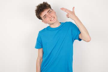 Fototapeta premium young caucasian handsome man with curly hair wearing blue T-shirt against white studio wall foolishes around shoots in temple with fingers makes suicide gesture. Funny model makes finger gun pistol