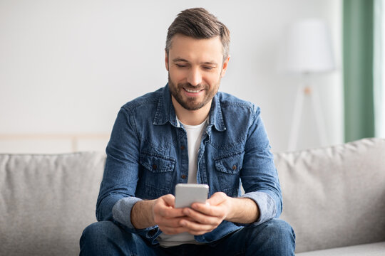 Happy Man Sitting On Couch At Home, Using Modern Smartphone