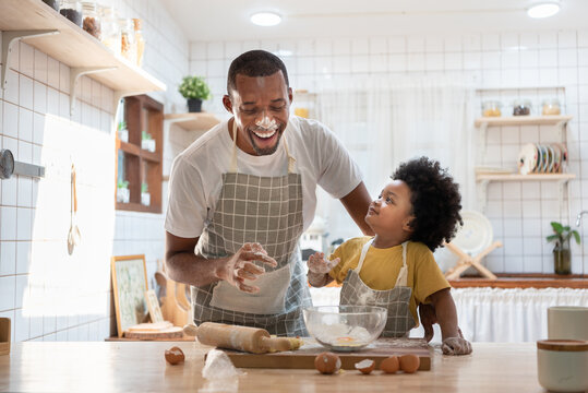 Cheerful smiling Black son enjoying playing with his father while doing bakery at home. Playful African family having fun cooking baking cake or cookies in kitchen together.