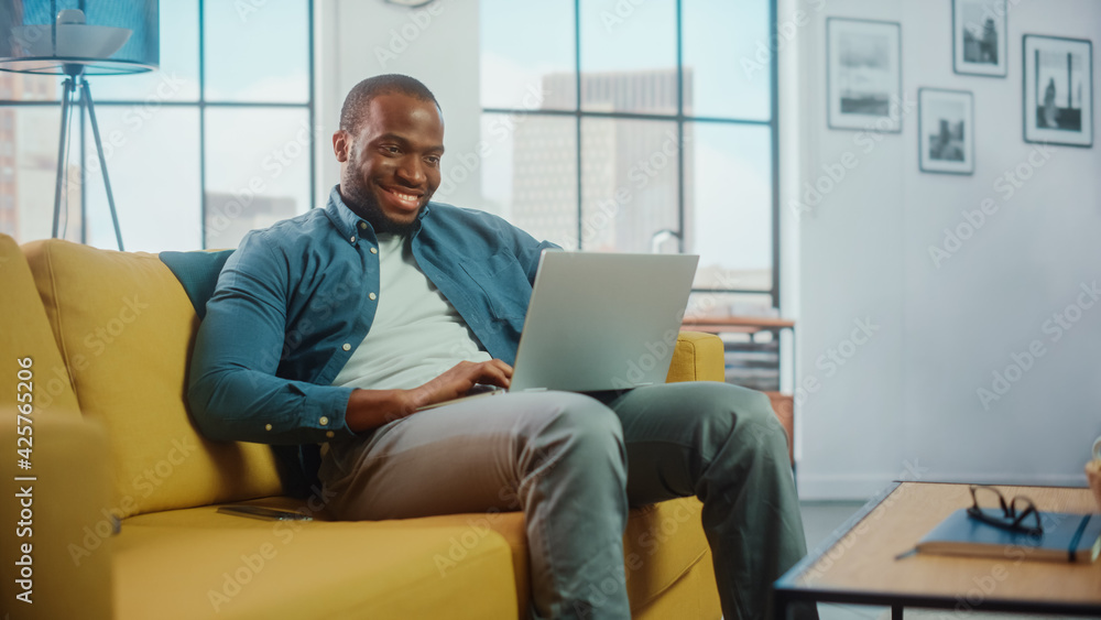 Poster handsome black african american man working on laptop computer while sitting on a sofa in cozy livin