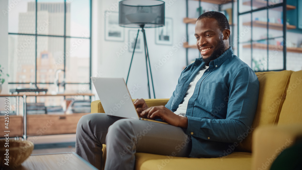 Poster handsome black african american man working on laptop computer while sitting on a sofa in cozy livin