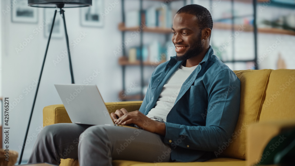 Poster handsome black african american man working on laptop computer while sitting on a sofa in cozy livin