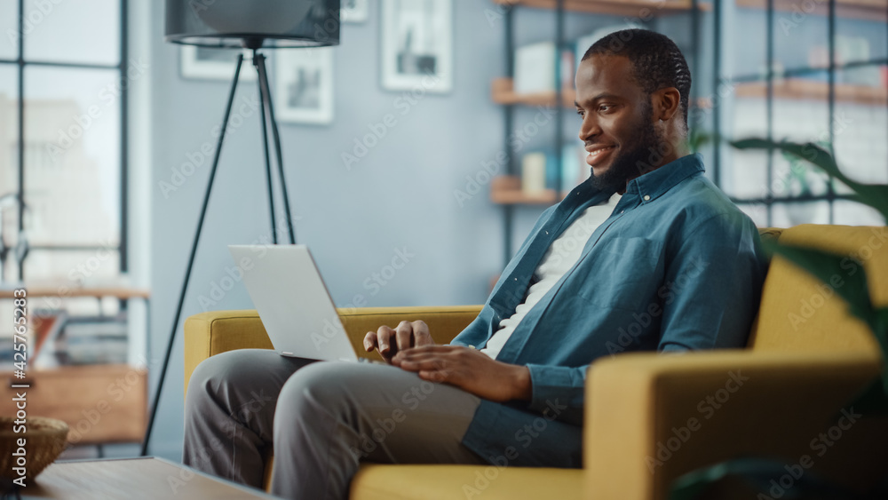 Poster handsome black african american man working on laptop computer while sitting on a sofa in cozy livin