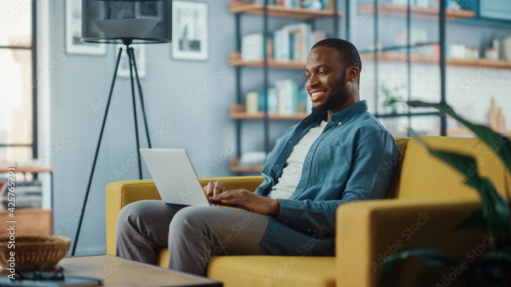 Poster handsome black african american man working on laptop computer while sitting on a sofa in cozy livin