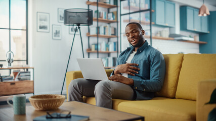 Handsome Black African American Man Having a Video Call on Laptop Computer while Sitting on a Sofa...