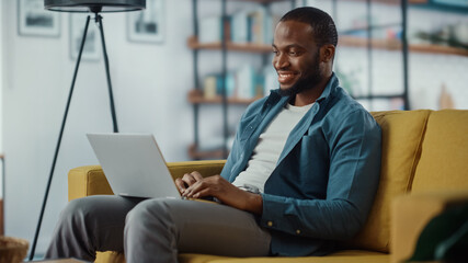 Handsome Black African American Man Working on Laptop Computer while Sitting on a Sofa in Cozy...